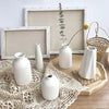 A collection of white ceramic vases and a dried plant displayed on a wooden surface with framed art.