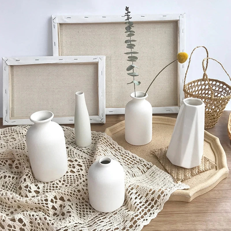A collection of white ceramic vases and a dried plant displayed on a wooden surface with framed art.