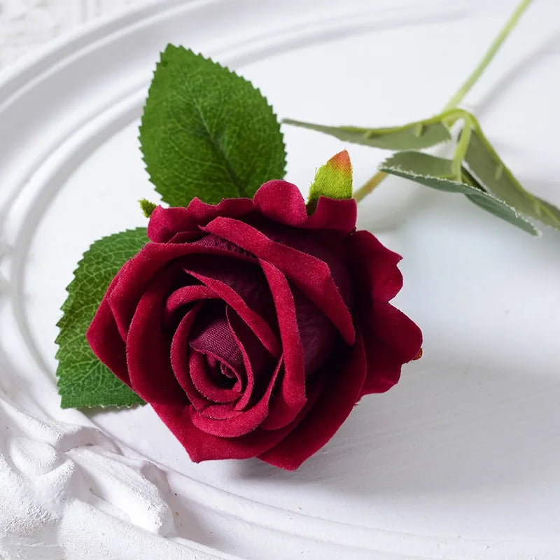 A deep red velvet artificial rose with green leaves and a bendable stem placed on a white plate.
