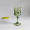 A clear green reusable plastic wine glass with a stem and foot sits on a white surface next to two lime halves.