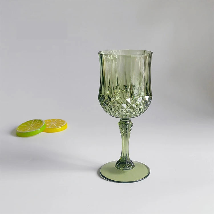 A clear green reusable plastic wine glass with a stem and foot sits on a white surface next to two lime halves.