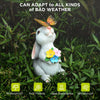 A white rabbit garden statue with a butterfly, surrounded by green foliage and rain.