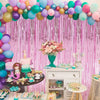 A vibrant pink and purple balloon backdrop decorated with metallic foil tassels and floral arrangements for a party.