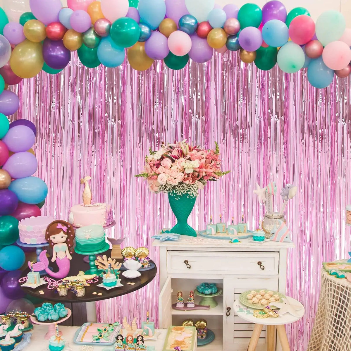 A vibrant pink and purple balloon backdrop decorated with metallic foil tassels and floral arrangements for a party.