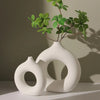 White ceramic vase shaped like a donut holding green foliage.