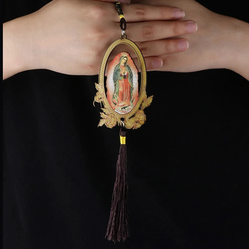 A wooden Our Lady of Guadalupe pendant charm with a gold-colored oval frame and a dark tassel, held in a person's hand.
