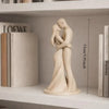 A minimalist sculpture of a couple hugging, made of light beige stone or ceramic, displayed on a white shelf.