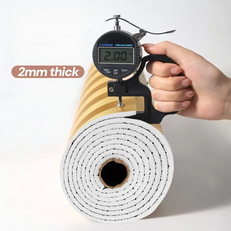 A hand holds a digital thickness gauge measuring 2mm thick metallic gold striped peel-and-stick wallpaper rolled on a spool.