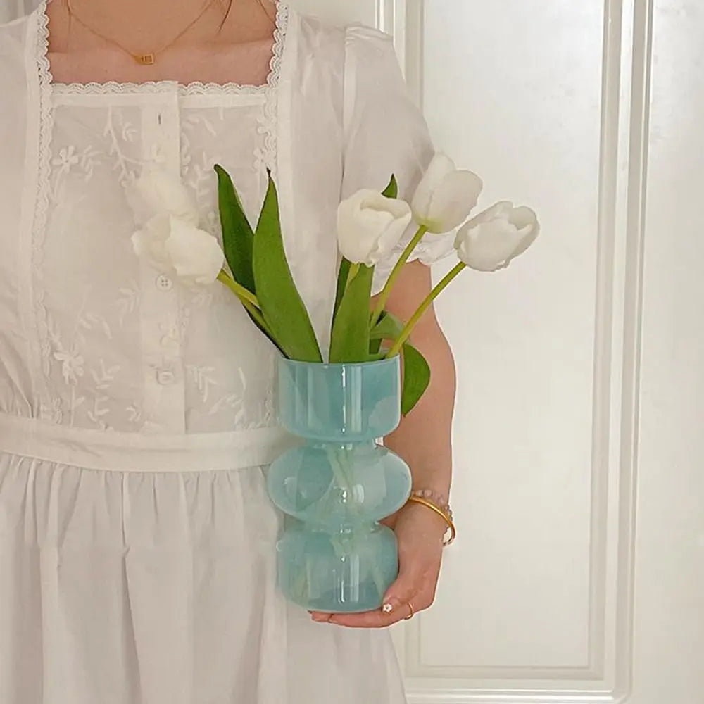 A person holds a light blue glass flower vase filled with white tulips against a white background.