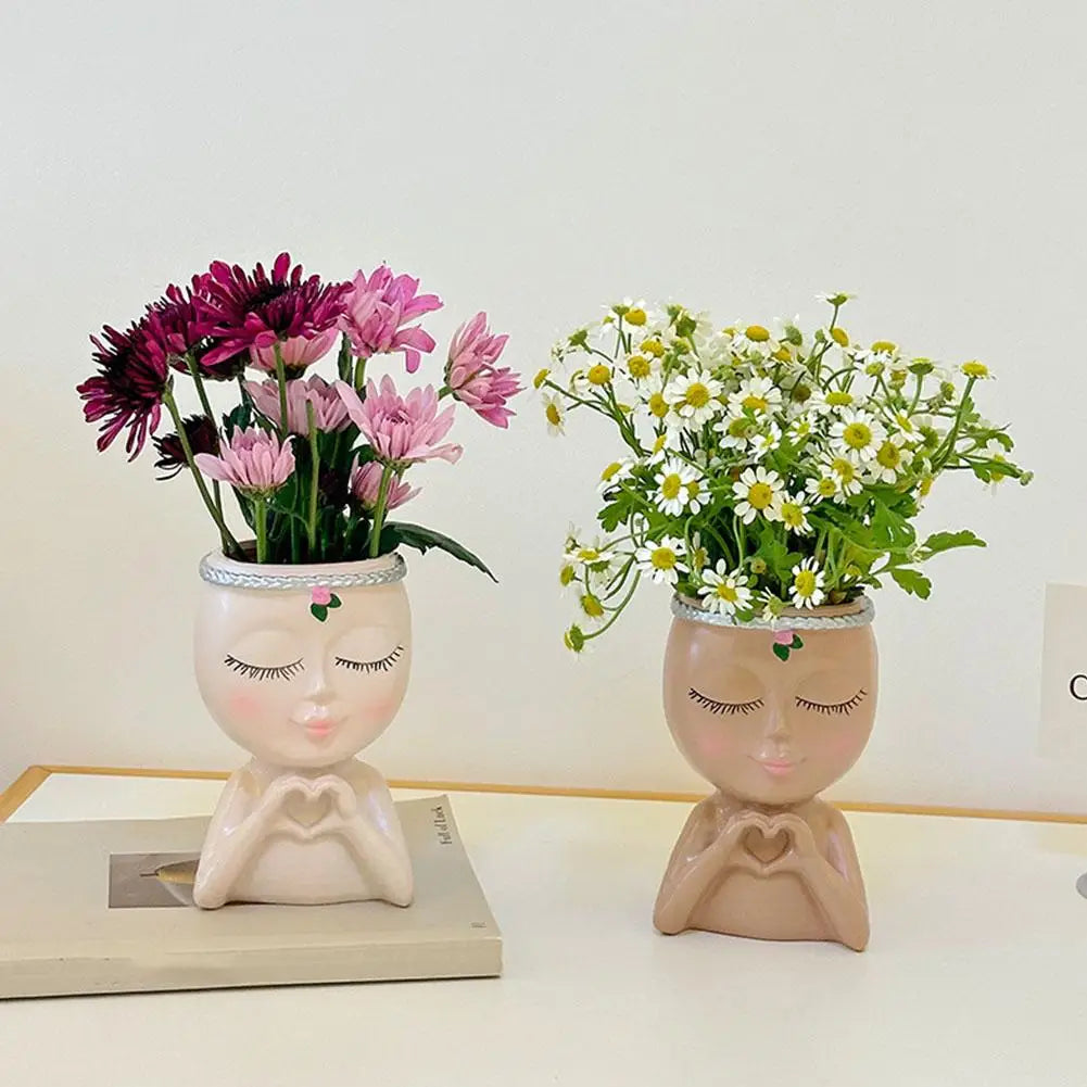 A pair of artistic resin girl face planters holding vibrant pink and purple flowers.