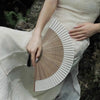 A hand holds a classic bamboo folding fan with a decorative pendant, displayed against a light fabric background.