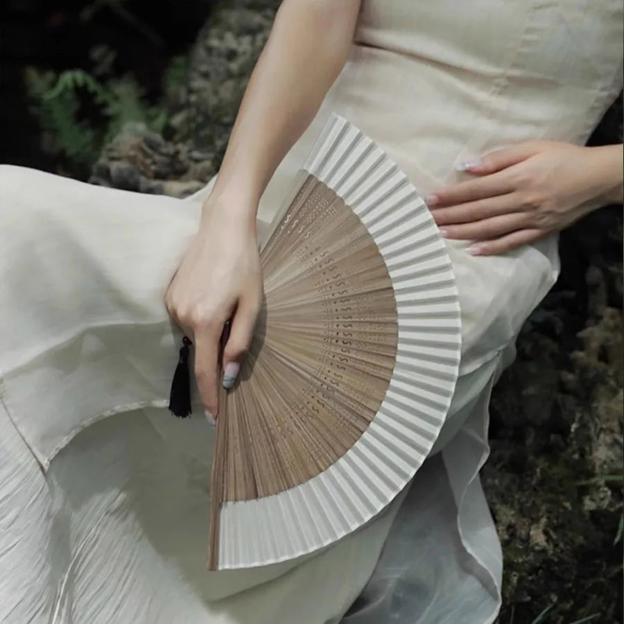 A hand holds a classic bamboo folding fan with a decorative pendant, displayed against a light fabric background.