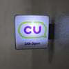 A rectangular sign with a purple CU logo inside a speech bubble and the text 24h Open, mounted on a wall.