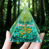Hand holding a clear resin pyramid filled with green stones and a copper coil in a forest setting