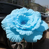 A large, light blue artificial peony flower is displayed on the hood of a dark car.