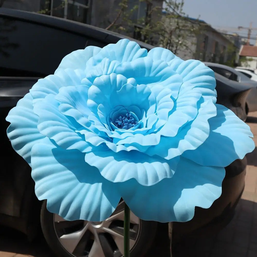 A large, light blue artificial peony flower is displayed on the hood of a dark car.