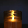 A glowing pillar candle in a glass holder with a flame visible inside, set against a dark background.