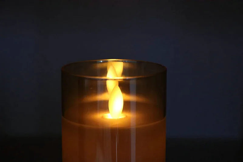 A glowing pillar candle in a glass holder with a flame visible inside, set against a dark background.