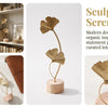 A golden ginkgo leaf ornament on a slender stem displayed on a round wooden base.