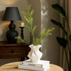 A white ceramic vase with a wave pattern holds green foliage on a wooden table next to a lamp.
