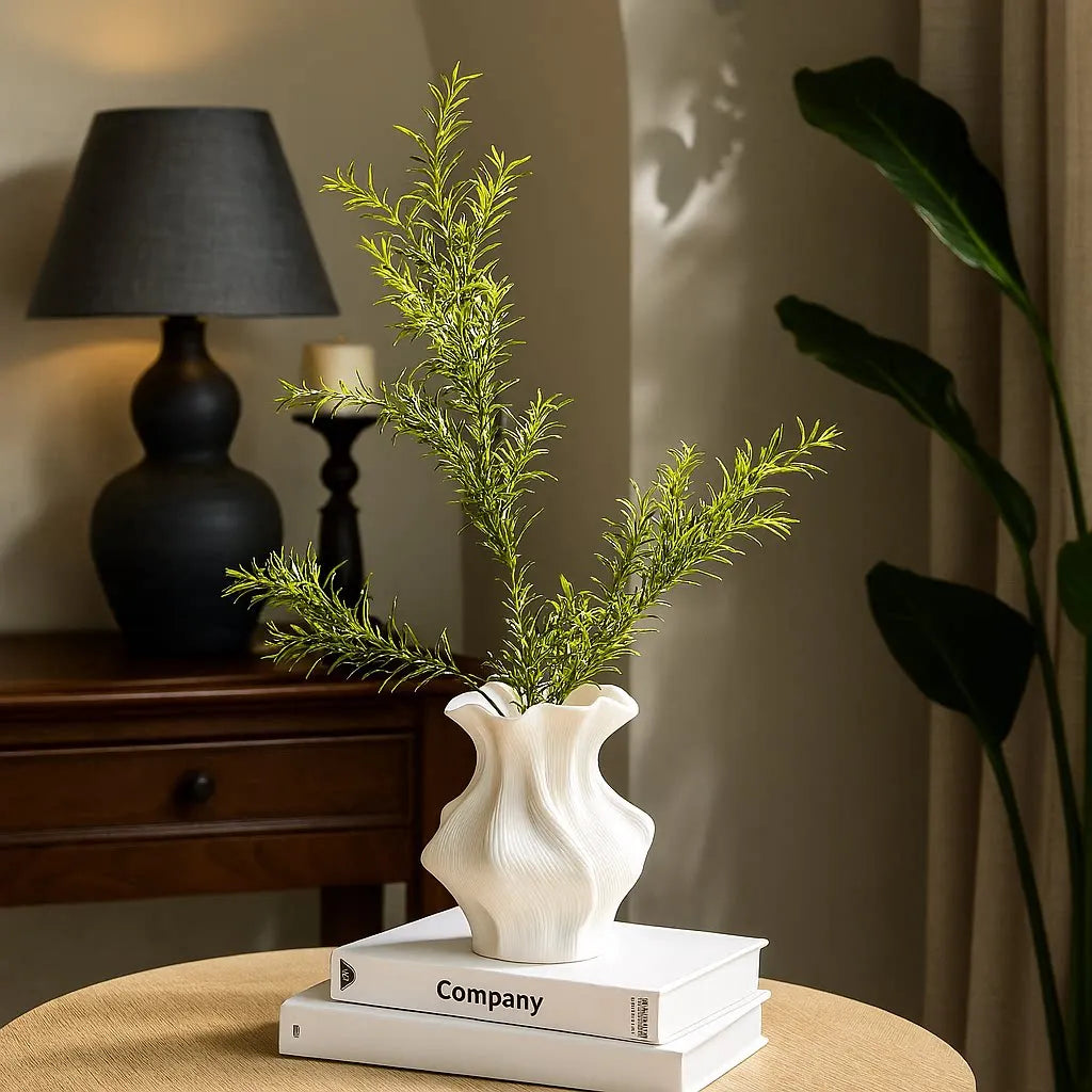 A white ceramic vase with a wave pattern holds green foliage on a wooden table next to a lamp.