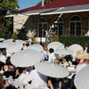 Guests at an outdoor event holding white decorative paper parasols under a covered structure.