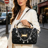 A woman models a black quilted handbag with gold chain and charms against an urban street background.