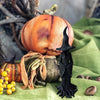 Estatua esbelta de bruja oscura de material negro junto a una calabaza naranja y follaje seco sobre tela verde.