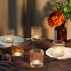 A collection of clear glass votive candle holders and an orange rose in a dark glass vase set on a brown fabric.