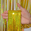 A hand holds a vertical strand of bright yellow metallic foil tinsel fringe against a shimmering gold and white backdrop.