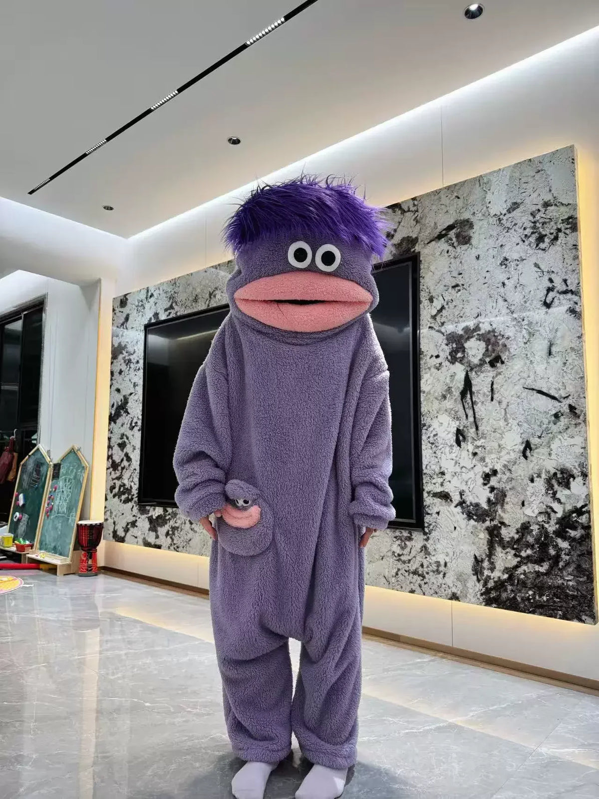 A purple monster onesie pajama suit with large eyes and a pink mouth stands in a brightly lit room.