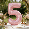 A large pink foil number balloon shaped like the number 5 with a bow, set in an outdoor floral and greenery context.