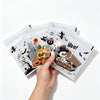 A hand holds several clear self-adhesive treat bags decorated with Halloween themes like bats skulls and pumpkins.