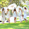 Five women in white dresses hold open white oil paper umbrellas outdoors on a sunny lawn.