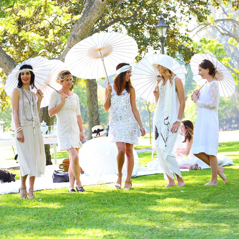 Five women in white dresses hold open white oil paper umbrellas outdoors on a sunny lawn.
