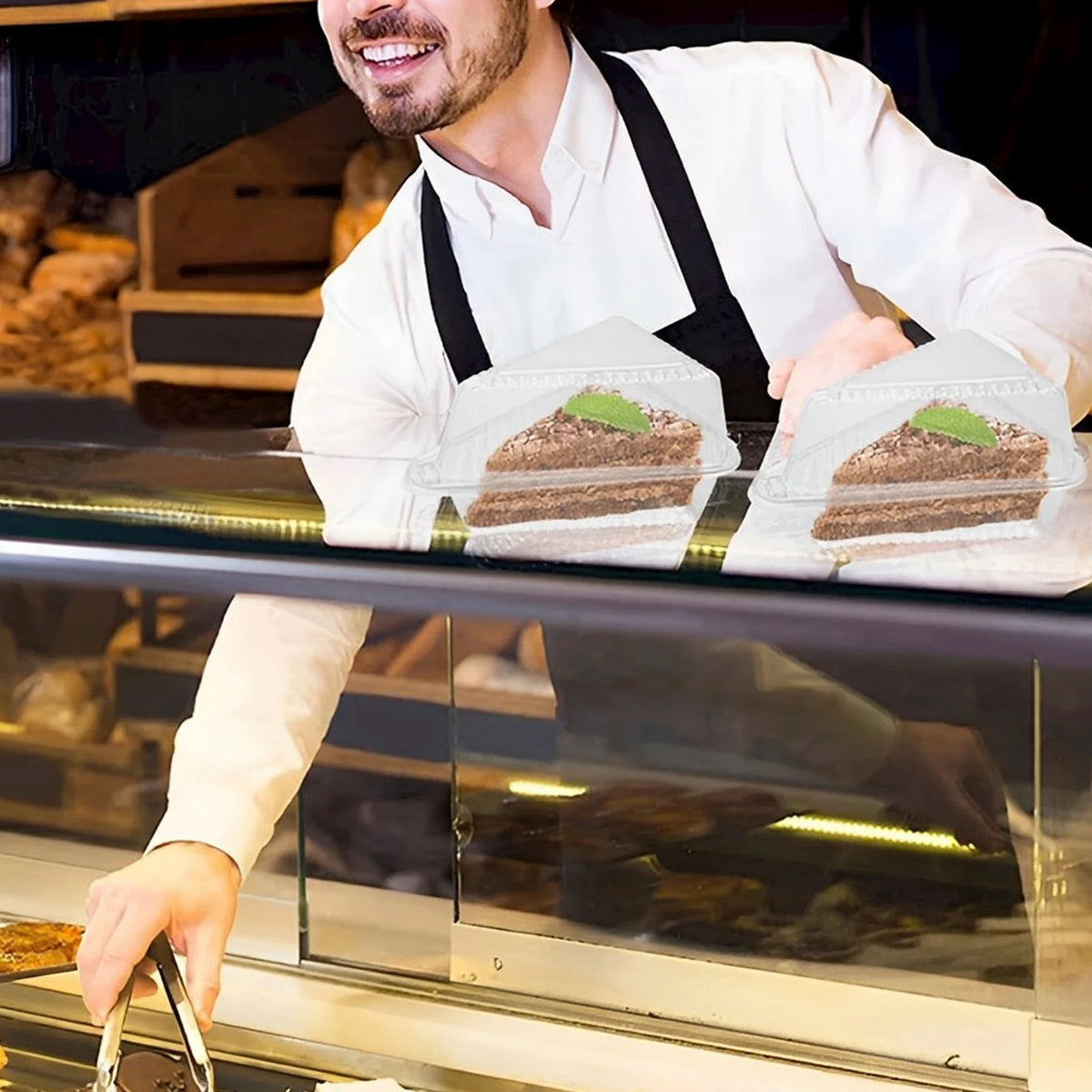 A man in a white shirt and apron is preparing or handling clear triangle cake slice containers filled with cake.