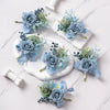 A set of six dusty blue silk rose boutonnières featuring delicate greenery and small blue filler flowers on white marble.