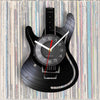 A black guitar-shaped wall clock with a vinyl record face and hands, set against a background of stacked vinyl records.