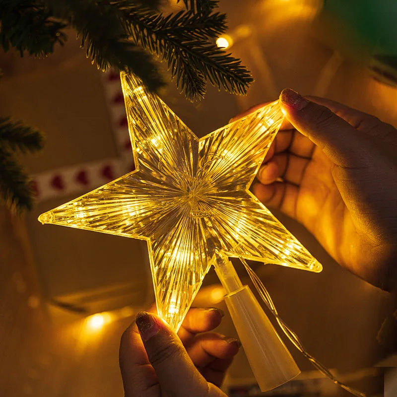 A hand holds a glowing LED star shaped Christmas tree topper with bright yellow light against a dark background.