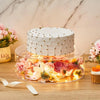 A tiered white cake decorated with gold accents sits on a clear acrylic stand filled with colorful dried flowers and illumina