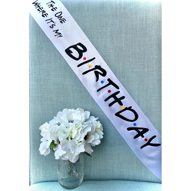 A white satin birthday sash with black lettering that reads Where It's My Birthday laid across a light blue fabric background