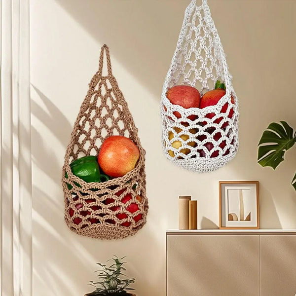 A macrame hanging storage basket filled with fresh fruits and vegetables on a light-colored wall.