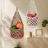A macrame hanging storage basket filled with fresh fruits and vegetables on a light-colored wall.