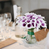 A bouquet of six artificial white calla lilies with purple accents in a glass vase on a table.