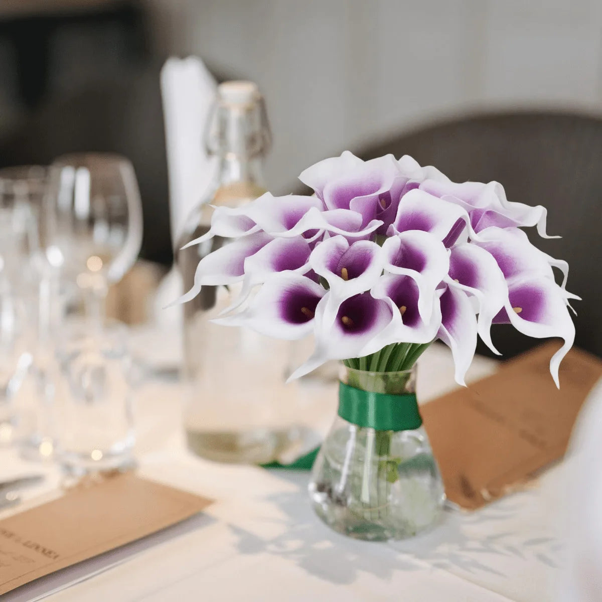 A bouquet of six artificial white calla lilies with purple accents in a glass vase on a table.