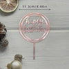 A pink acrylic birthday cake topper with gold lettering and a thin stick, set against a light gray wooden background.