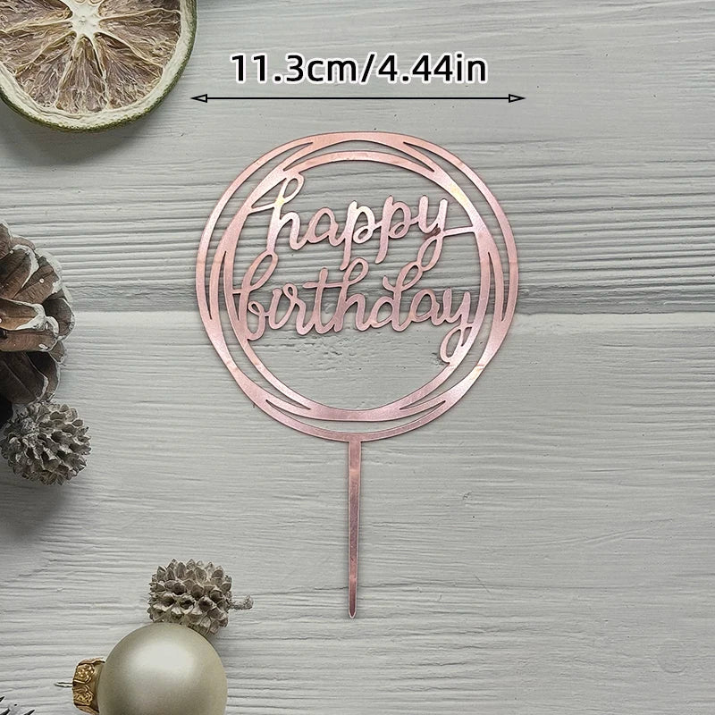 A pink acrylic birthday cake topper with gold lettering and a thin stick, set against a light gray wooden background.