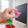 A hand holds a pink dinosaur fridge magnet against a dark chalkboard background.