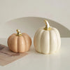 A white ceramic pumpkin figurine with a gold stem sits on a light surface next to a pink-toned pumpkin.