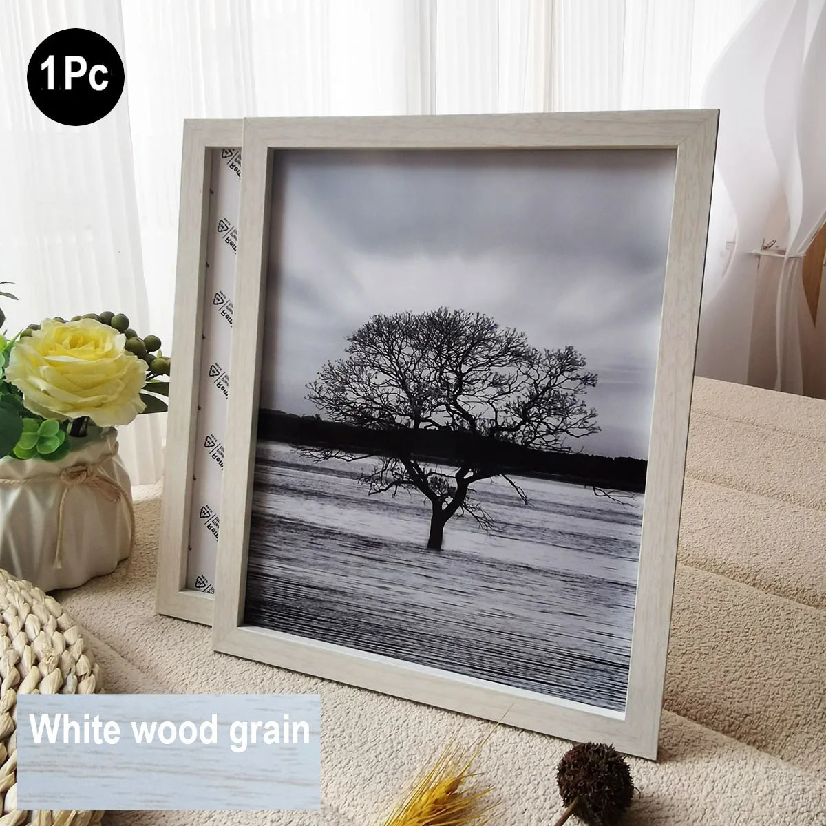 A framed black and white photograph of a bare tree against a cloudy sky, displayed in a white wood grain frame.